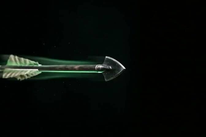 A broadhead arrow frozen mid-flight, trailing a green streak through the dark.